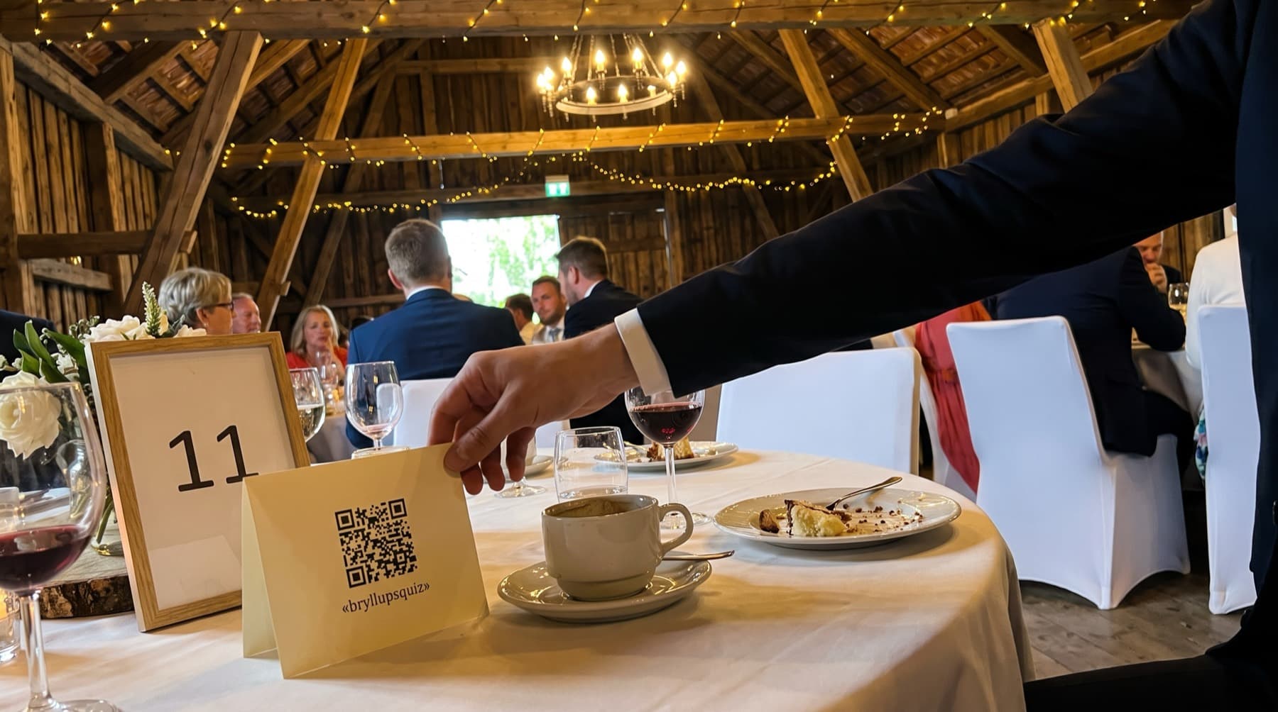 Et lite kort med ordet bryllupsquiz står ved bordnummeret på et rundt bord, ved siden av en kaffekopp og en kakeasjett. En hånd tar det.